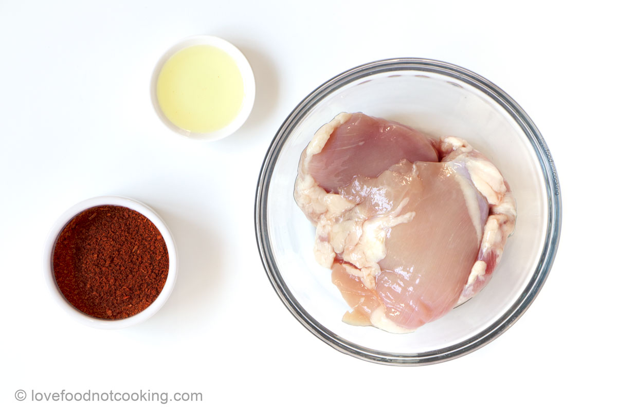 Uncooked chicken thighs, seasoning and oil in separate bowls on a white background. 