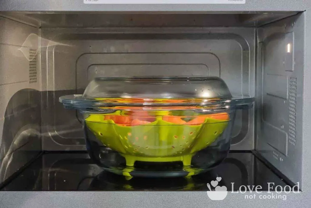 glass bowl containing green microwave steamer basket and carrots in microwave. 