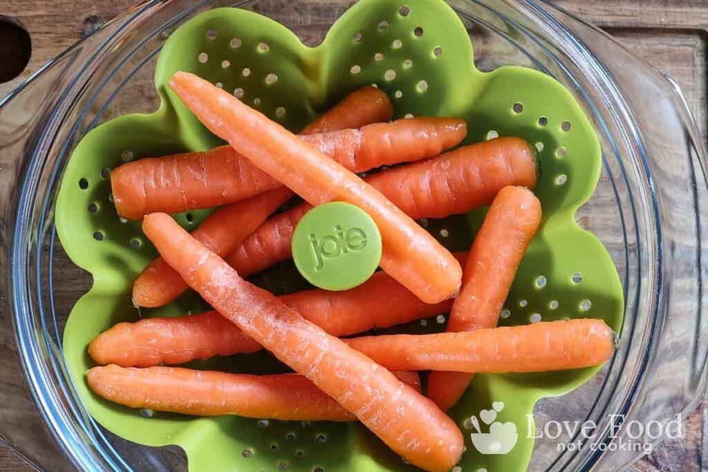 Baby carrots in a green steamer basket in a glass bowl. 