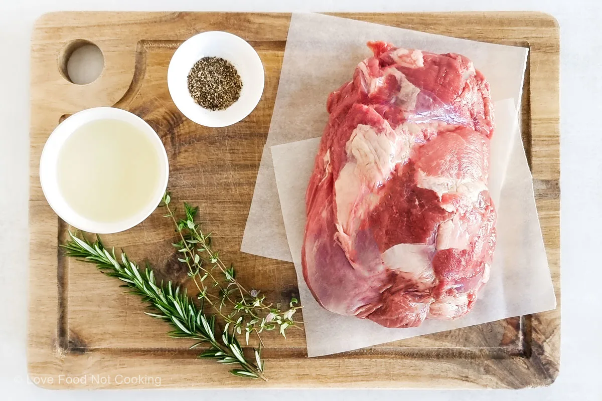 Boneless lamb leg roast on a wooden board with oil, black pepper, rosemary and thyme.