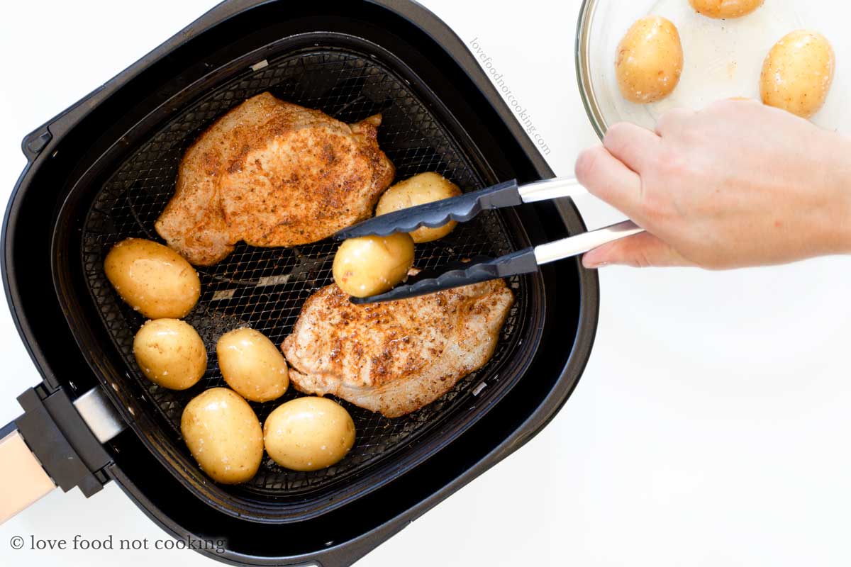 Pork chops and potatoes in air fryer basket, potatoes being added by hand. 