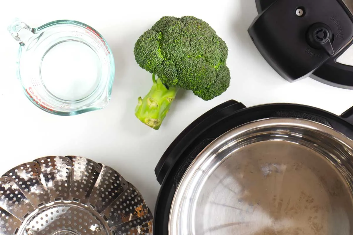 An Instant Pot, a steamer basket, a jug of water, and a head of broccoli. 