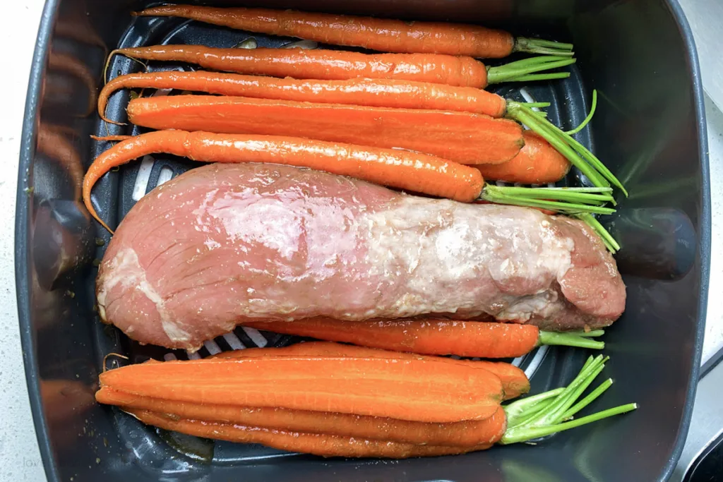 Uncooked pork tenderloin in air fryer basket with carrots.