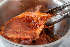Pork chop held in tongs coated in seasoning and oil.